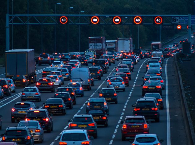 Image shows a traffic jam, it is evening so there are brake lights and the speed signs are illuminated on the overhead bridge