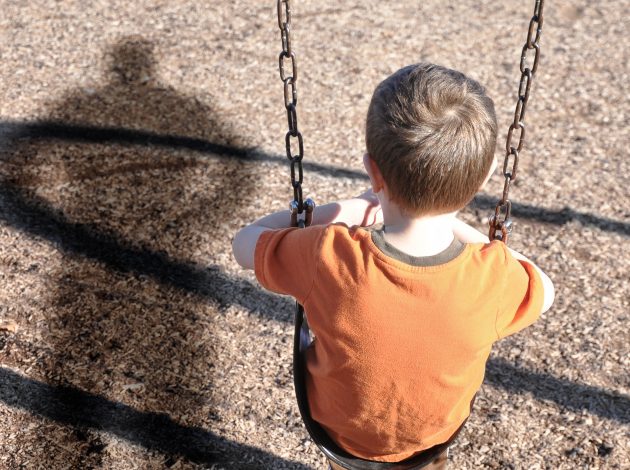 A young boy is sitting on a swing set and looking at a shadow figure of a man or bully at a playground