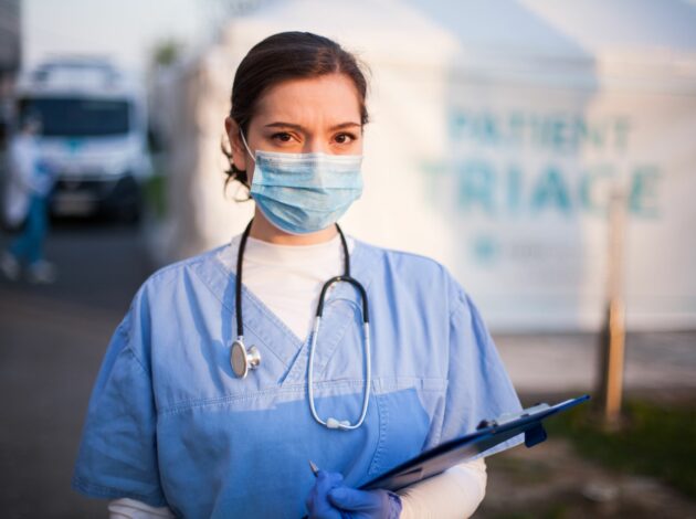 An image of a woman wearing surgical scrubs and a mask. She has a stethoscope around her neck and is holding a clipboard.