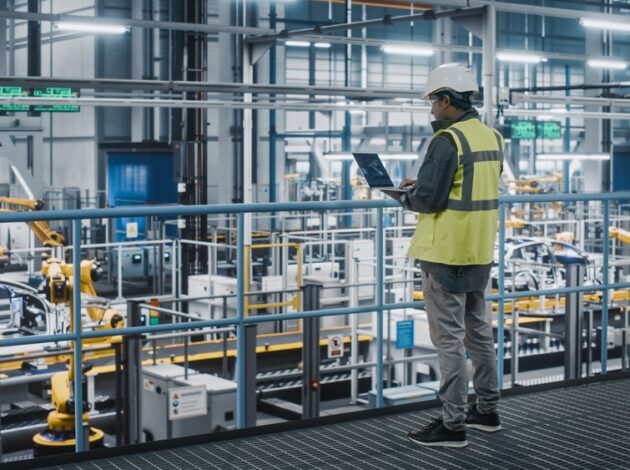 Car Factory Engineer in High Visibility Vest Using Laptop Computer in an Automotive Industrial Manufacturing Facility